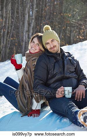 Stock Photography - Portrait of couple sitting in snow with hot drinks. Fotosearch