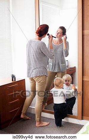 Stock Image - Woman applying make up with son at mirror. Fotosearch