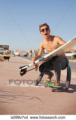 Stock Image - Young man skateboarding down street whilst holding a surfboard. Fotosearch