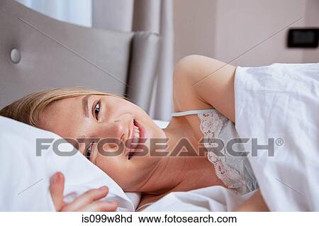 Stock Image - Young woman lying in bed and smiling at camera. Fotosearch