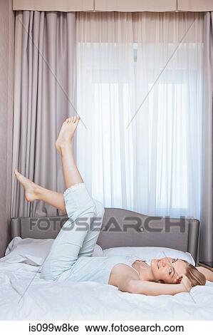 Stock Image - Young woman lying on bed with legs in the air. Fotosearch