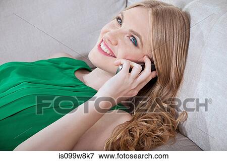 Stock Photo - Young woman on cellphone, lying on sofa. Fotosearch