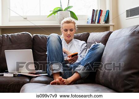 Stock Photo - Young woman sitting on sofa and looking at smartphone. Fotosearch