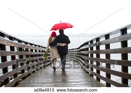 Stock Image - Couple walking on wooden pier in rain. Fotosearch