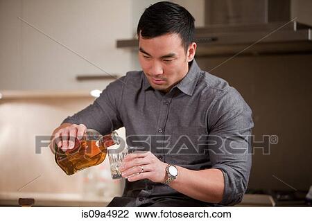 Stock Image - Man pouring glass of whiskey in kitchen. Fotosearch
