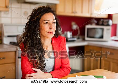 Stock Image - Smiling woman sitting in kitchen. Fotosearch