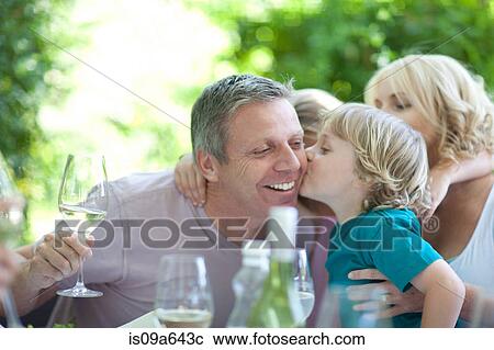 Stock Image - Boy kissing father at table outdoors. Fotosearch