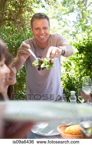 Stock Photograph - Man serving salad at table outdoors. Fotosearch
