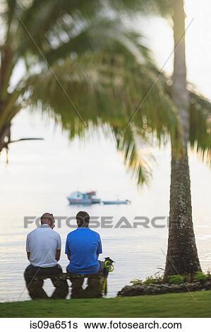 Stock Photograph - Men sitting under palm tree on beach. Fotosearch