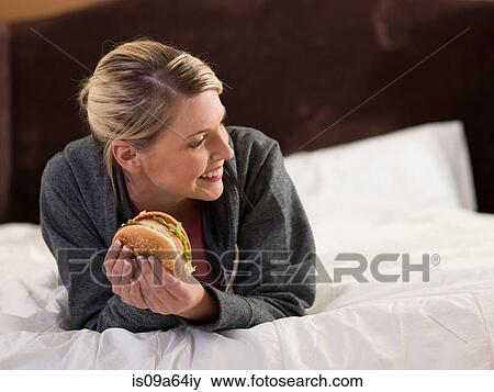 Stock Photo - Woman eating hamburger on bed. Fotosearch