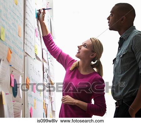 Stock Photo - Colleagues looking at adhesive notes and paper on wall. Fotosearch