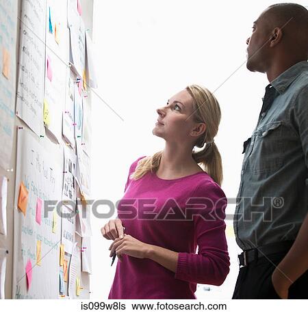 Colleagues looking at adhesive notes and paper on wall View Large Photo Image Stock Photograph - Colleagues looking at adhesive notes and paper on wall. Fotosearch