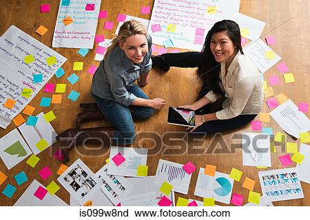 Stock Image - Colleagues sitting on floor with digital tablet, papers and adhesive notes. Fotosearch