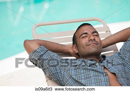 Stock Image - Man relaxing in lawn chair by pool. Fotosearch