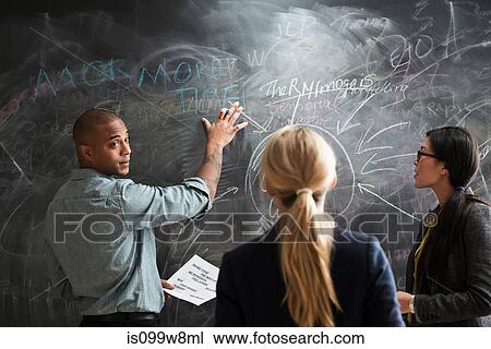 Stock Image - Man writing on blackboard, colleagues watching. Fotosearch