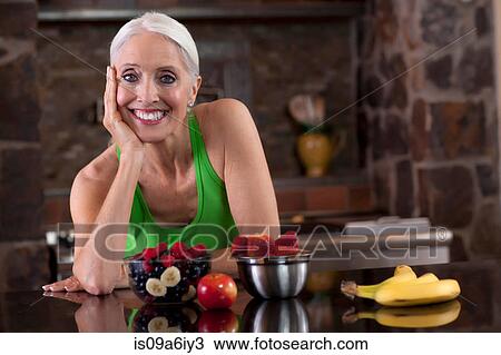 Stock Image - Older woman having fruit in kitchen. Fotosearch