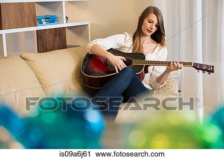 Stock Image - Woman playing guitar on sofa. Fotosearch