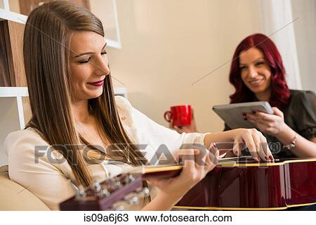 Stock Image - Woman playing guitar on sofa. Fotosearch