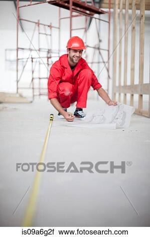 Stock Image - Builder with blue print measuring floor on construction site. Fotosearch
