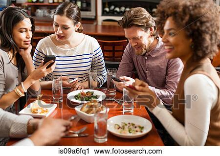 Stock Image - Friends at restaurant texting and showing photos using cell phones. Fotosearch