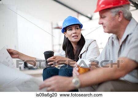 Stock Image - Male and female construction workers sitting down talking and holding coffee and a blueprint. Fotosearch