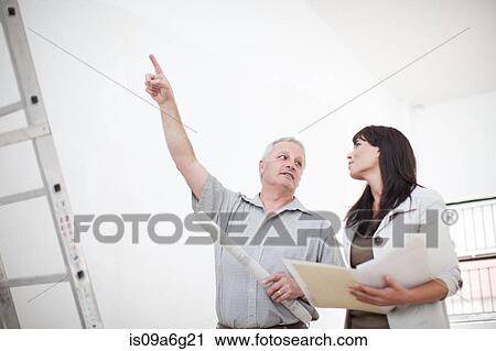 Stock Image - Male and female talking and pointing upwards holding blueprints next to ladders. Fotosearch