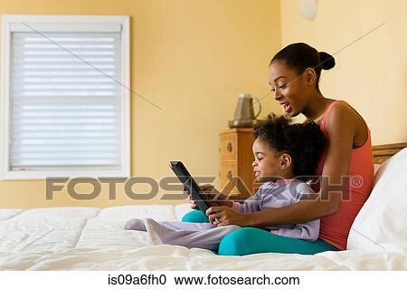 Stock Image - Mother and daughter sitting on bed using digital tablet. Fotosearch