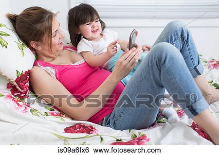 Portrait of pregnant woman and toddler daughter lounging on bed playing with smartphone View Large Photo Image Stock Photograph - Portrait of pregnant woman and toddler daughter lounging on bed playing with smartphone. Fotosearch