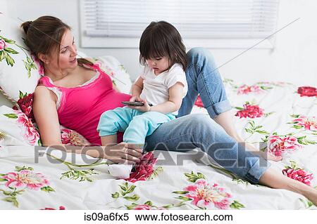 Stock Photography - Portrait of pregnant woman and toddler daughter lounging on bed with smartphone. Fotosearch