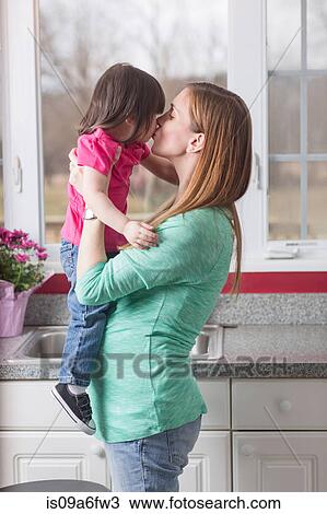 Stock Image - Pregnant mother holding toddler daughter in kitchen. Fotosearch