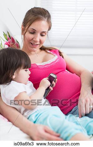 Stock Image - Pregnant woman and toddler daughter looking at smartphone. Fotosearch