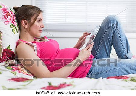 Stock Image - Pregnant woman lying on bed looking at digital tablet. Fotosearch