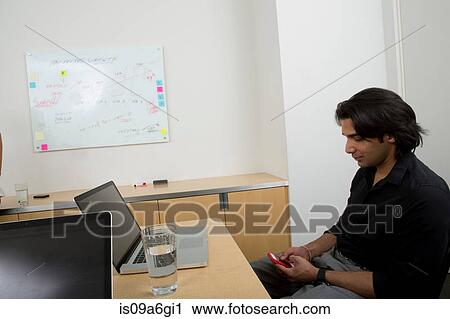 Young man at desk with cell phone View Large Photo Image Stock Image - Young man at desk with cell phone. Fotosearch