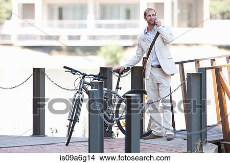 Picture - Young man by chain fence with bicycle on cell phone. Fotosearch