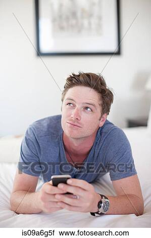 Stock Image - Young man holding smartphone, looking away. Fotosearch