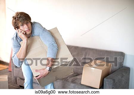 Stock Image - Young man moving house on cell phone carrying box. Fotosearch