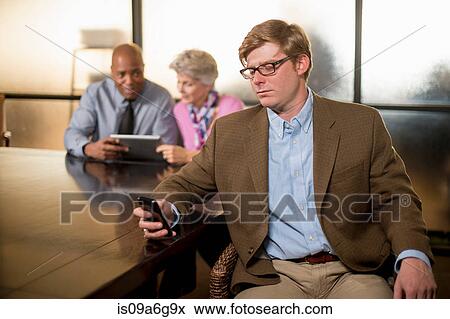 Stock Photo - Young man using cell phone with colleagues in background. Fotosearch