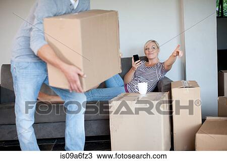 Stock Image - Young woman moving house on sofa directing man with box. Fotosearch