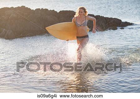 Stock Image - Young woman running in sea with surfboard. Fotosearch