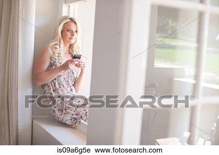Young woman sitting on windowsill holding smartphone View Large Photo Image Stock Image - Young woman sitting on windowsill holding smartphone. Fotosearch