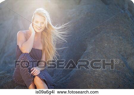 Stock Image - Young woman with long blonde hair sitting on rocks. Fotosearch