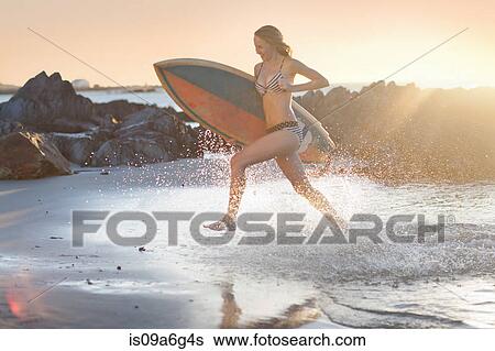 Stock Photograph - Young woman with surfboard running on beach. Fotosearch