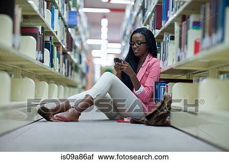 Stock Image - Female student sitting on floor in library using smartphone. Fotosearch