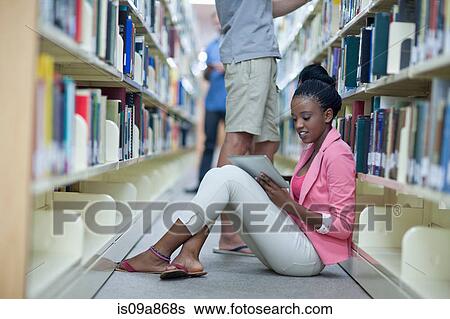 Stock Photograph - Female student sitting on floor in library using digital tablet. Fotosearch