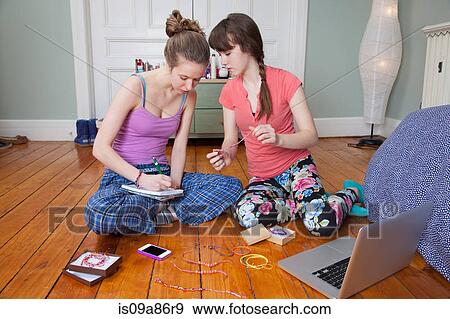 Stock Photo - Girls sitting on bedroom floor with laptop, making friendship bracelets. Fotosearch