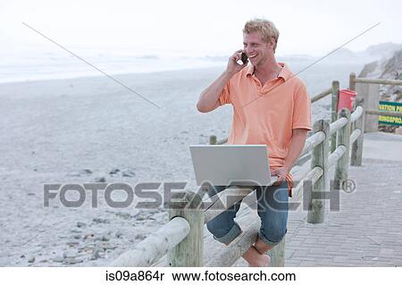 Stock Photography - Man sitting on beach railing using phone and laptop. Fotosearch