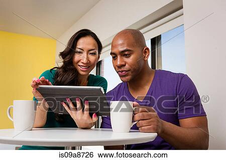 Stock Photograph - Mid adult couple looking at digital tablet and drinking coffee. Fotosearch