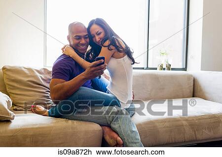 Stock Image - Mid adult couple together on sofa looking at smartphone. Fotosearch