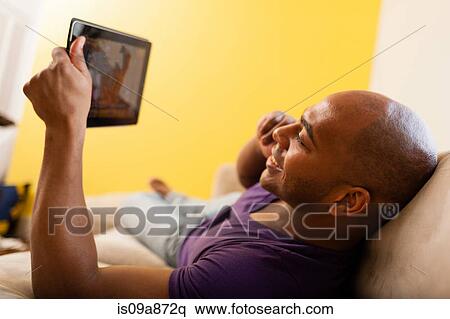 Stock Photography - Mid adult male on sofa holding digital tablet and mobile phone. Fotosearch