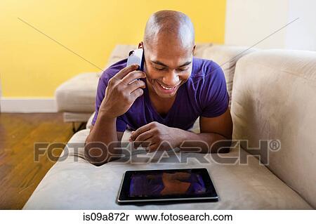 Stock Photography - Mid adult male on sofa using digital tablet and smartphone. Fotosearch